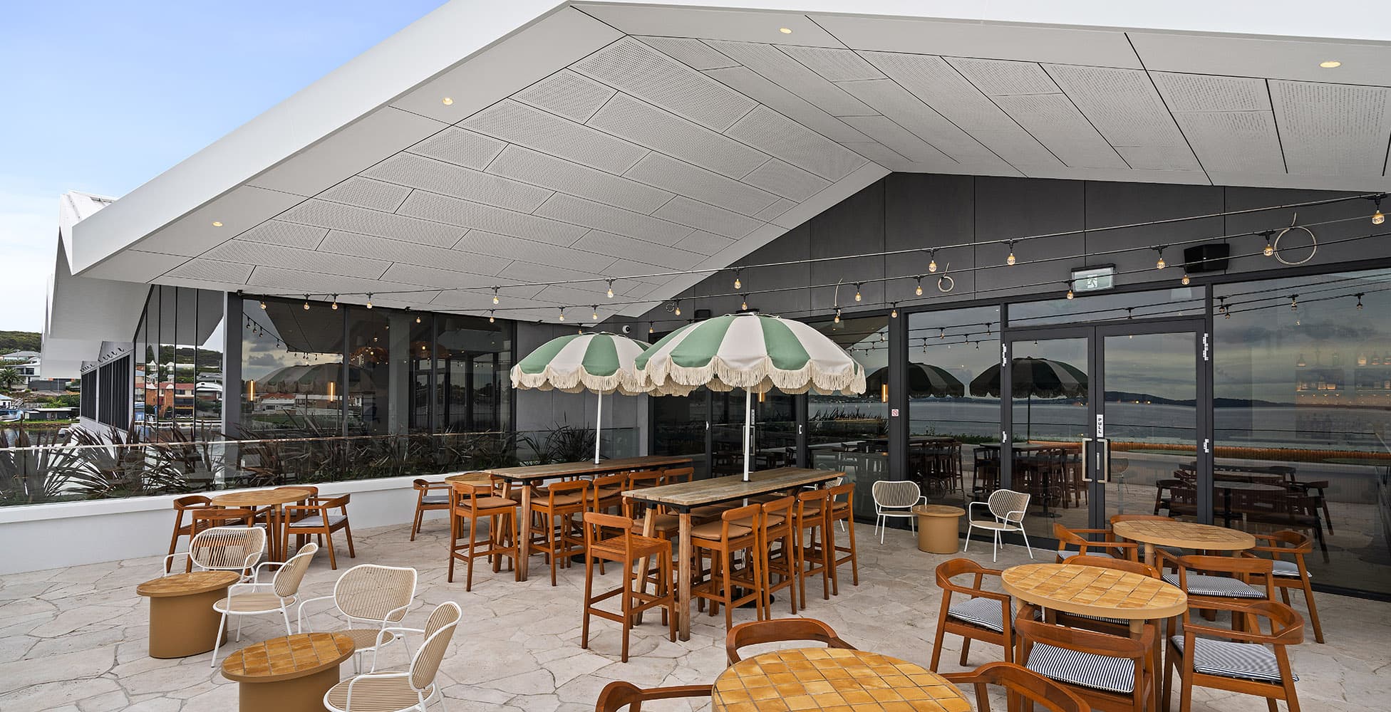 The rooftop terrace at Belmont 16s with outdoor dining furniture under a dramatic angular canopy roof, green-and-white striped umbrellas, and views across Lake Macquarie.