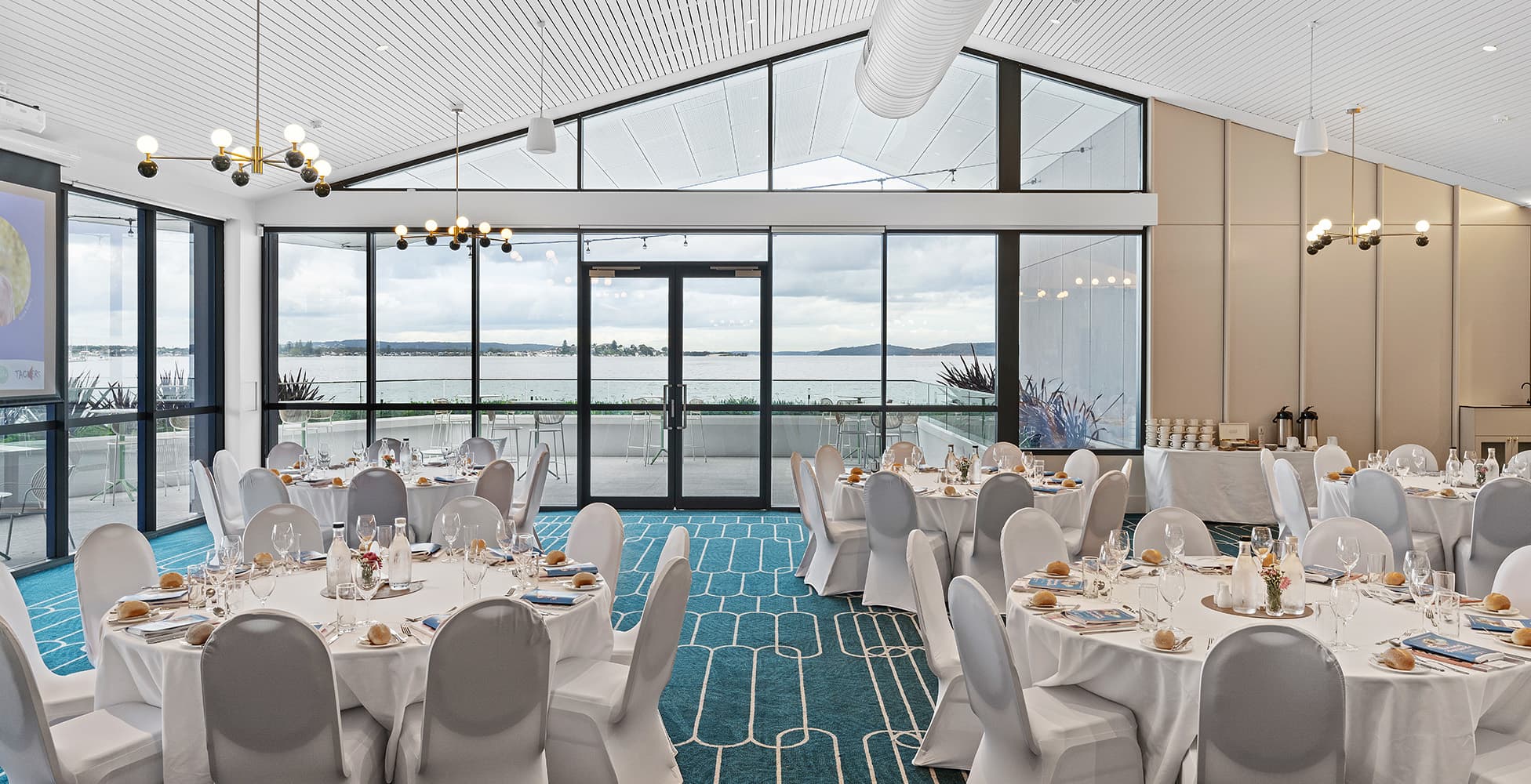 The new event and function space at Belmont 16s with panoramic lake views through floor-to-ceiling glazed walls, a dramatic white pitched ceiling, modern chandeliers, and formal banquet table settings.