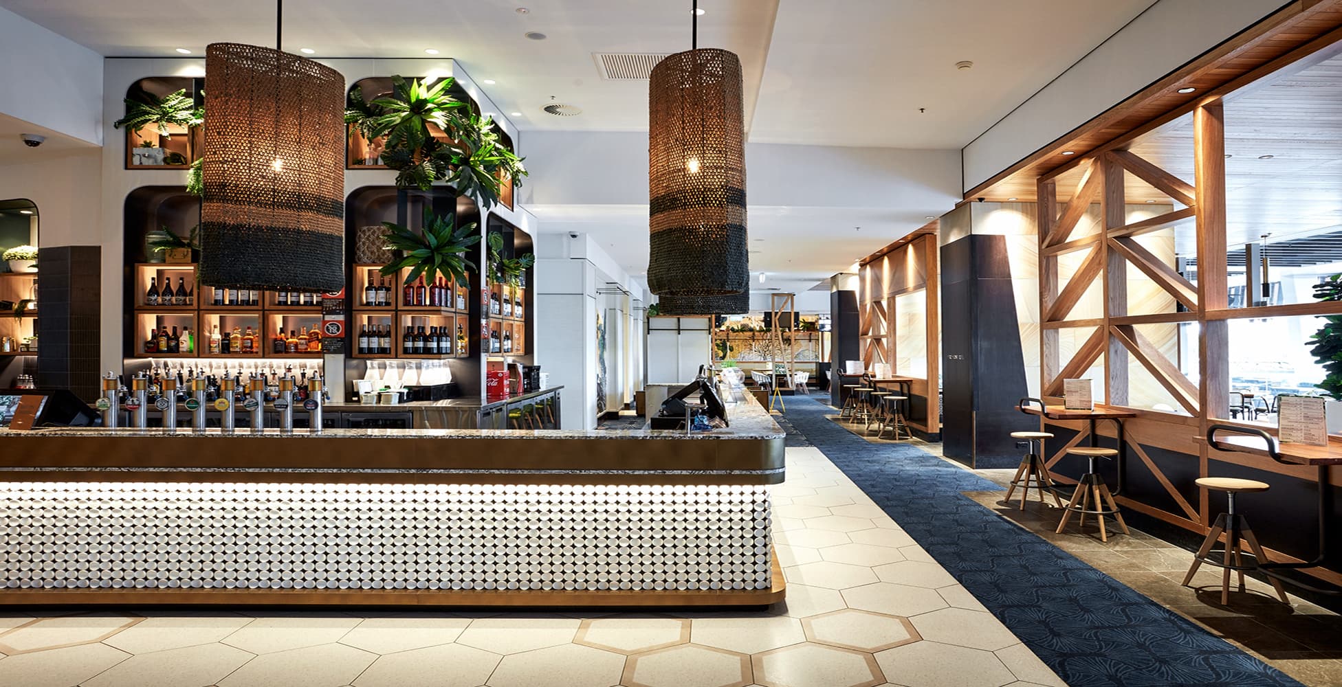 The main bar at Canterbury League Club, featuring wicker pendant lights, tropical greenery, white tile bar front, and a warm coastal-inspired material palette with timber and stone detailing.