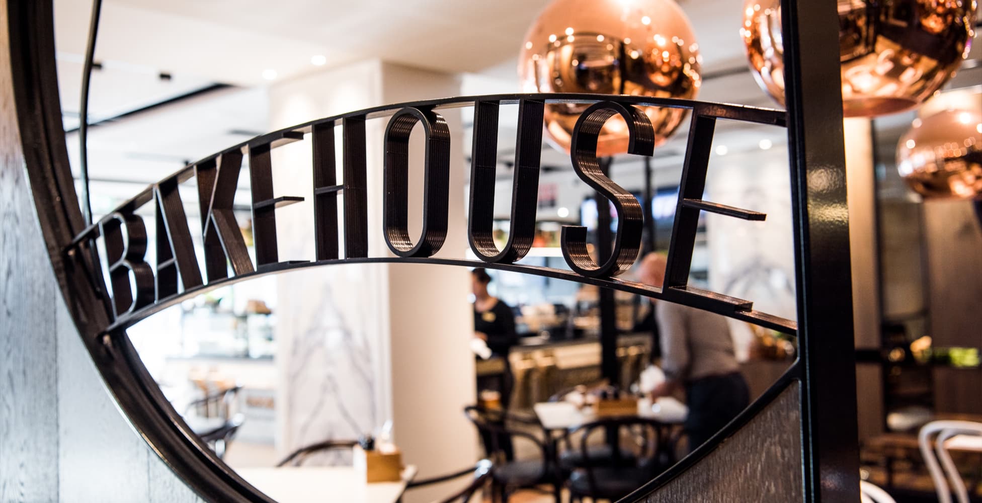 The Bakehouse cafe entrance at Canterbury League Club, showing the signature wrought-iron 'Bakehouse' arch, copper pendant globes, and the art-nouveau inspired interior beyond.