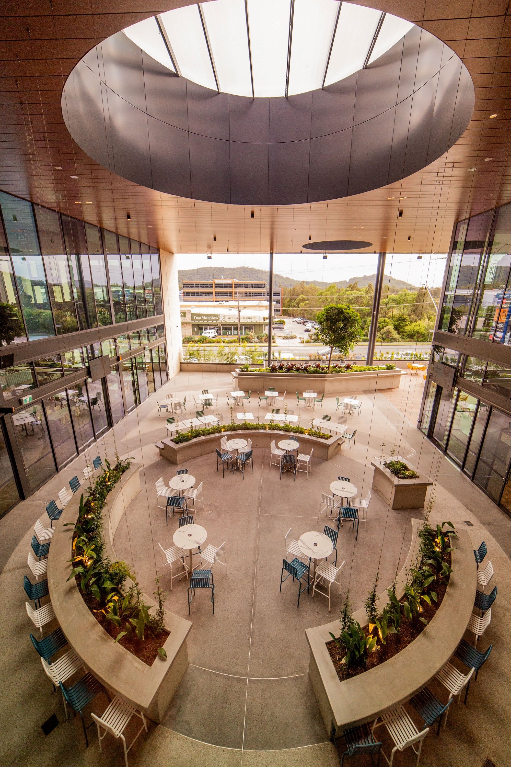 The double-height outdoor terrace viewed from above, showing a dramatic circular oculus skylight, timber-lined ceiling, lush planter boxes, and seating arranged around a curved garden setting with views to the surrounding hills.
