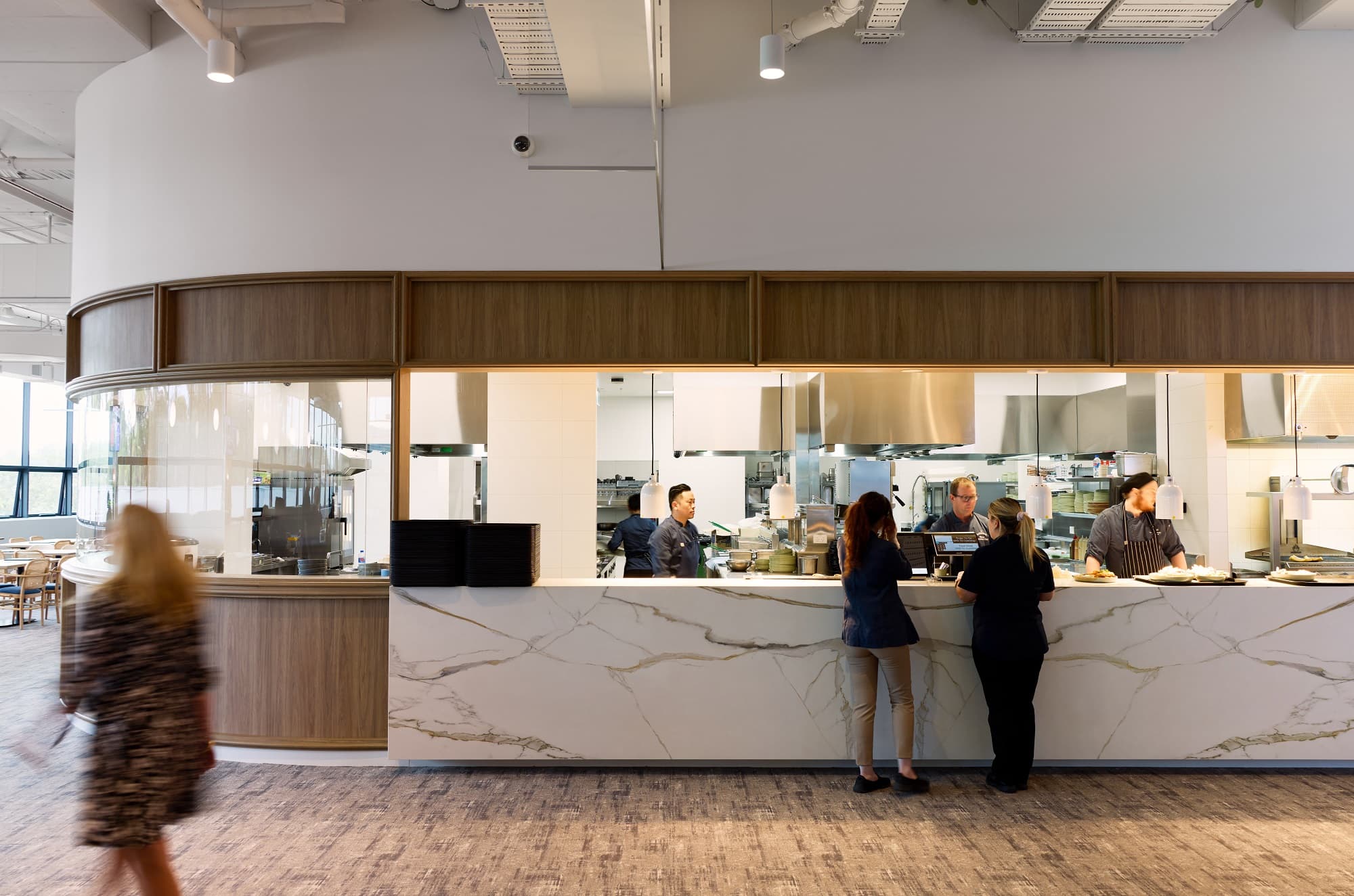 The open kitchen and dining area on Level 1, featuring a curved marble-effect service counter, timber-panelled walls with glass pass-through to the kitchen, and warm lighting throughout.