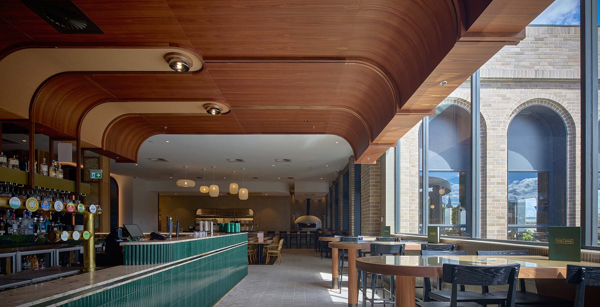 The upper-level bar and restaurant space featuring sweeping timber ceiling detail, arched windows with natural light, and dining tables overlooking the precinct.