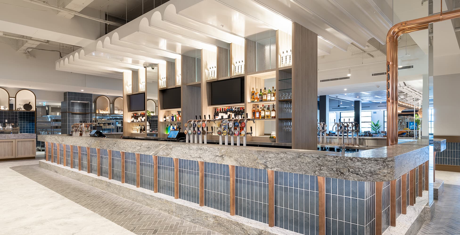 The central bar featuring copper pipe detailing, blue tile frontage with timber inserts, and a marble-effect benchtop with 80-plus beer taps.