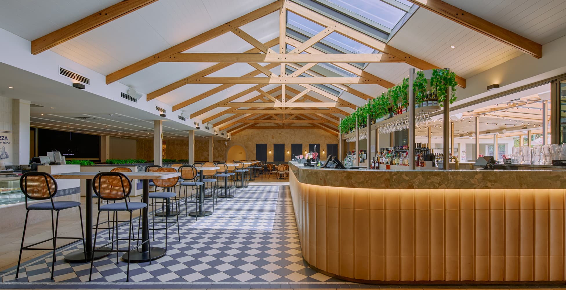 The main piazza bar area showing the full-length marble bar with leather upholstery, exposed timber roof trusses with skylights, blue-and-white checkerboard floor tiles, and high tables with bistro seating.