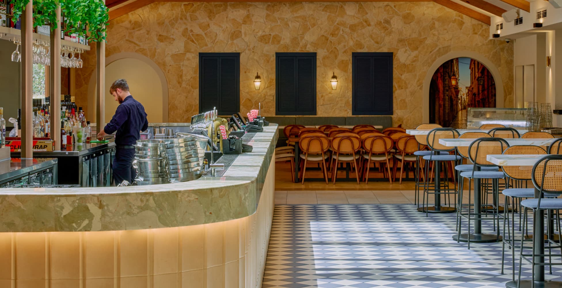 The stone-clad bar area featuring Croatian-inspired arched openings, shuttered windows, warm sandstone walls, green marble bar top, and a mix of dining and high-table seating on checkerboard tiles.