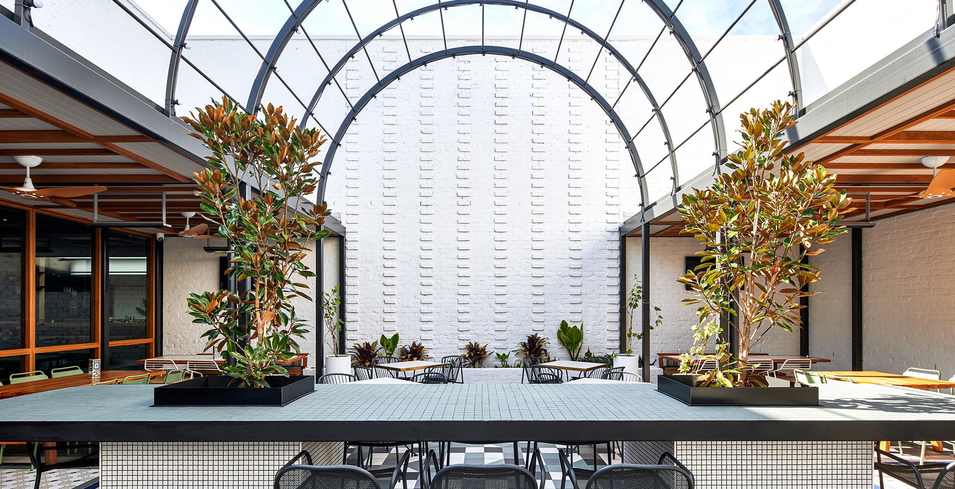The Atrium courtyard at Seven Hills RSL, featuring a dramatic arched glass canopy, mature plantings, and an outdoor bar — a standout feature of the Altis Architecture design.