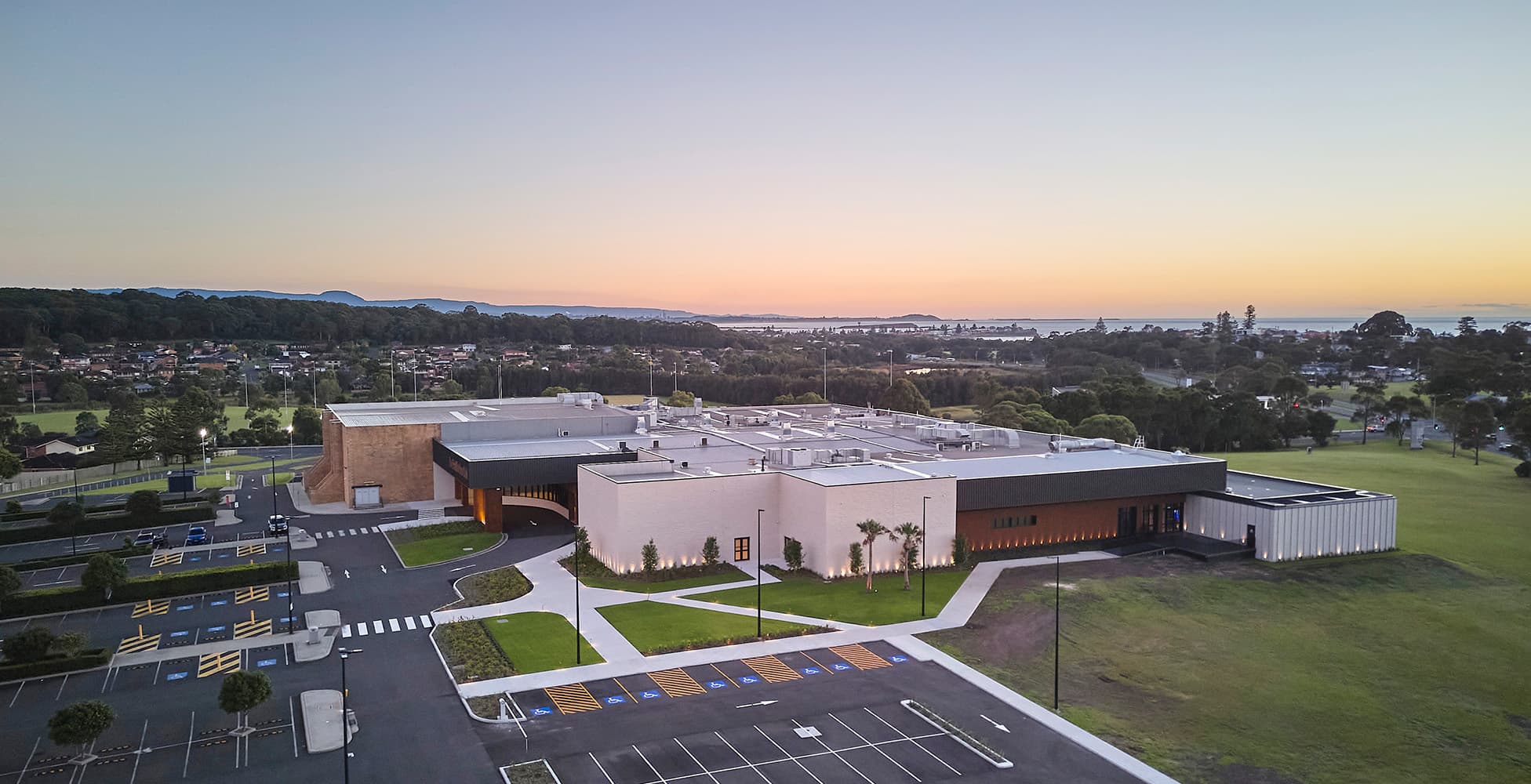 Aerial view of The Shellharbour Club showing the completed southern facade, new entry, landscaping, car park, and surrounding Illawarra landscape at dusk.