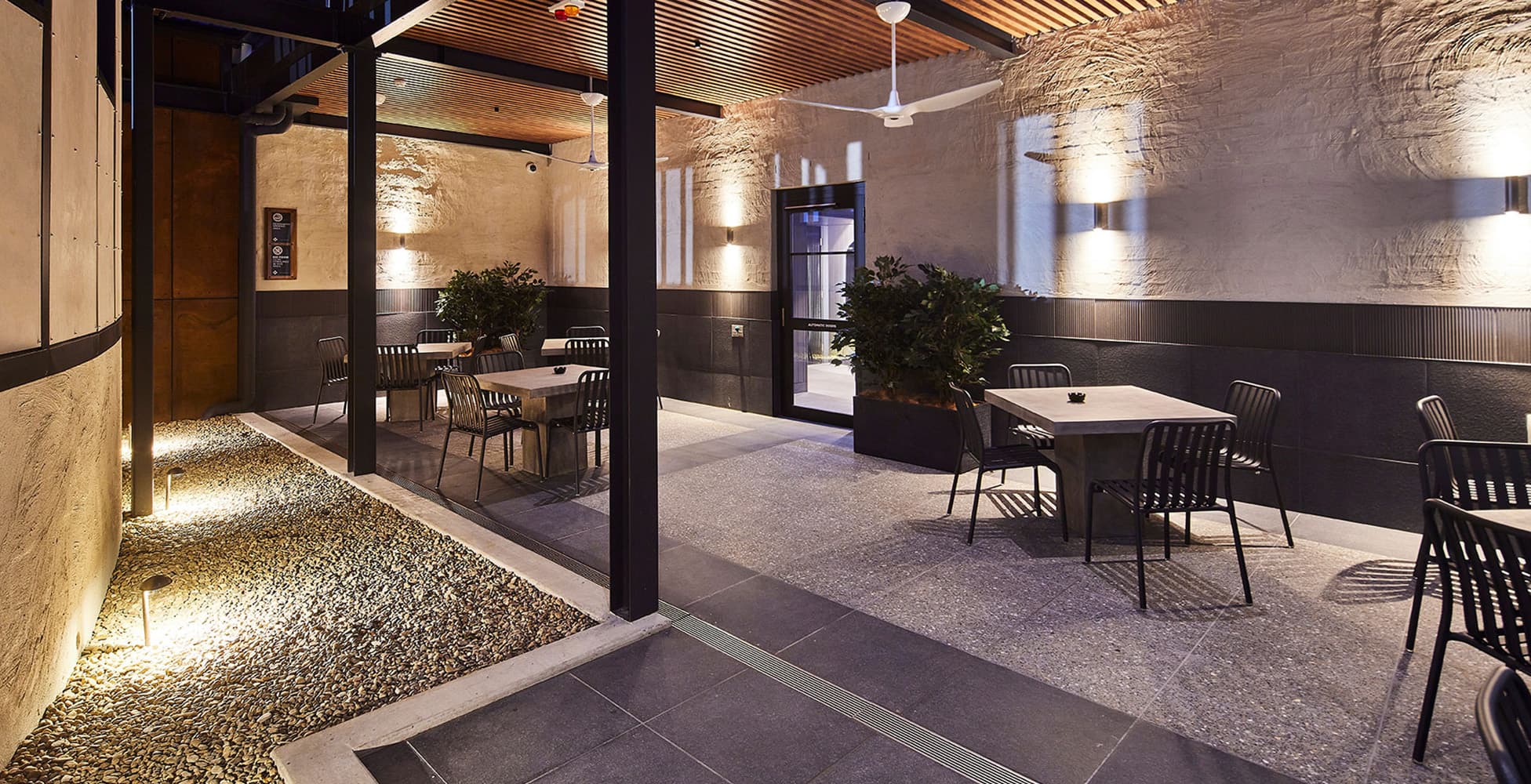 The outdoor terrace dining area at The Shellharbour Club, featuring raw concrete walls, timber-battened ceiling, ambient uplighting, and contemporary furniture.